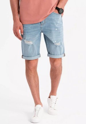 Shorts de mezclilla azul claro con detalles desgastados, dobladillos enrollados y salpicaduras de pintura blanca; combinados con zapatillas blancas.
