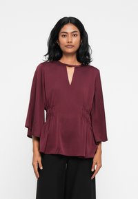 Blusa borgogna con scollatura a chiave, maniche a campana e arricciature laterali. Realizzata in un tessuto morbido, presenta una vestibilità comoda.