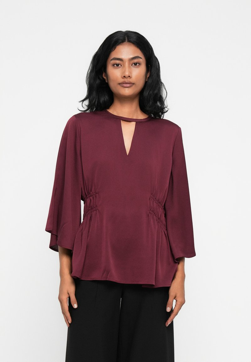 Blusa borgogna con scollatura a chiave, maniche a campana e arricciature laterali. Realizzata in un tessuto morbido, presenta una vestibilità comoda.