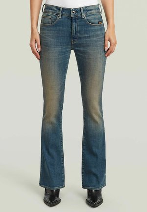 Blaue ausgewaschene Bootcut-Jeans, getragen mit schwarzen spitzen Schuhen, sichtbar an den Knöcheln.
