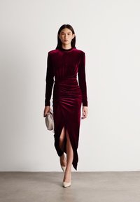 Βουργουνδί βελούδινο φόρεμα με μακριά μανίκια, ραμμένα λεπτομέρειες και υψηλό σκίσιμο. Συνδυασμένο με μπεζ ψηλές γόβες και μια μικρή ασημένια τσάντα.