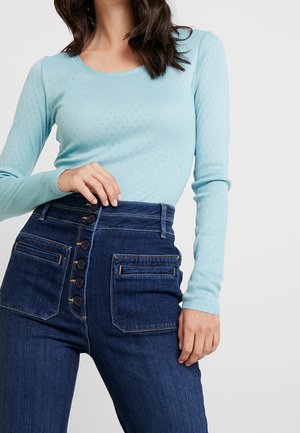 Mujer con top de punto de manga larga en azul claro y vaqueros de tiro alto azul oscuro con botones delanteros y grandes bolsillos.