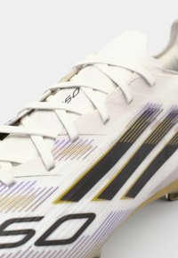 White athletic shoe with black and gold stripes, textured material, and intricate line patterns; features traditional lacing and reinforced toe.