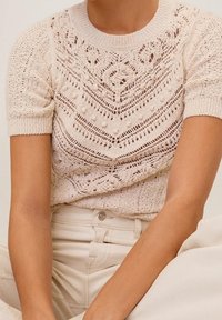 Krátký rukáv, pletený top v světlé béžové barvě s propracovanými průvlečnými vzory a texturovanými akcenty. Kulatý výstřih. Volný střih.