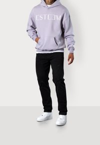 Felpa con cappuccio color lavanda con scritta "ESTEEM", tasca a marsupio, vestibilità comoda; abbinata a jeans neri strappati e sneakers bianche.