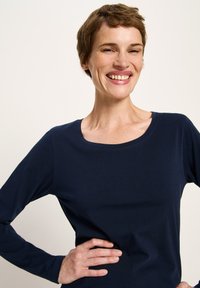 Lächelnde Frau mit kurzen braunen Haaren, die ein marineblaues Langarmshirt trägt und mit einer Hand auf der Hüfte vor einem schlichten Hintergrund steht.