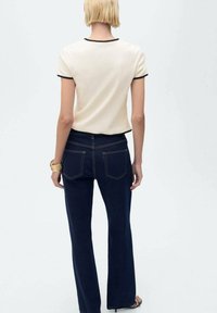 Haut beige à manches courtes avec bord noir, associé à un jean bleu foncé avec poches arrière et coupe droite. Texture lisse dans l'ensemble.