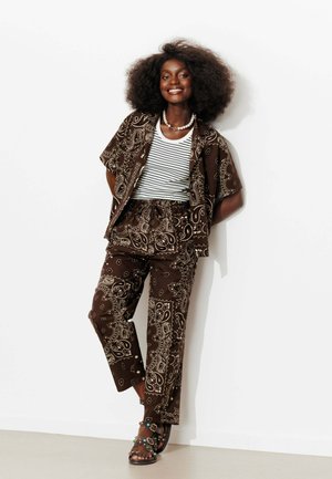 Femme aux grands cheveux bouclés sourit, appuyée contre un mur blanc, portant une chemise et un pantalon marron à motifs, un top rayé, des sandales et un collier de perles.