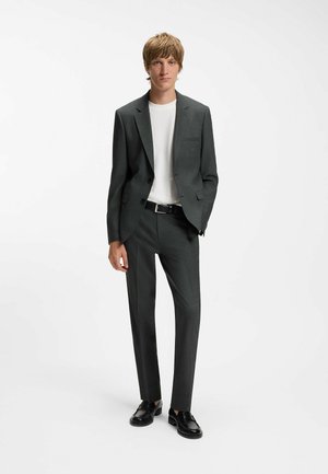 Jeune homme debout portant un costume gris foncé, un pull blanc, une ceinture noire et des chaussures habillées noires sur un fond clair uni.
