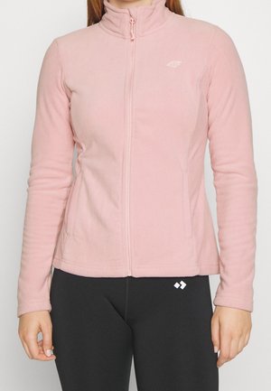 Femme portant une veste polaire rose clair zippée avec col montant et des leggings noirs avec un petit logo blanc.