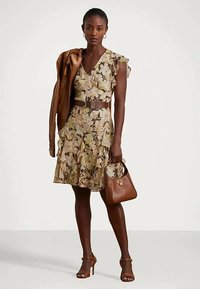 Moteris vilki gėlėtą suknelę su volanais ir ruda diržu, laiko rudą odinį švarką per petį ir suderintą rudą rankinę.