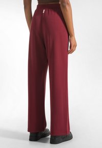 Bordeaux wijdbeenbroek met een gladde textuur, elastische tailleband en zwarte schoenen. Zichtbare stiksels en een klein labeldetail nabij de tailleband.