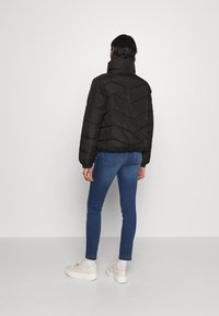 Giacca a vento nera con motivo a zigzag, colletto alto e polsini elastici, abbinata a jeans blu e sneakers color crema.