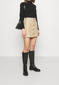 Beige Leder-Minirock mit vorderem Knopfverschluss, kombiniert mit einem schwarzen langärmeligen Oberteil, knielangen schwarzen Stiefeln und einer kleinen, gesteppte schwarzen Tasche.
