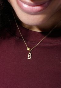 Goldene Halskette mit einem Anhänger in Blockbuchstaben "B" und einem kleinen Charm, der an einer zarten Kette vor einem bordeauxfarbenen Stoffhintergrund hängt.