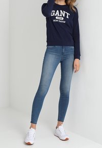 Sweatshirt blu navy con logo "GANT", abbinato a jeans skinny blu chiaro e sneaker bianche con tocchi arancioni.