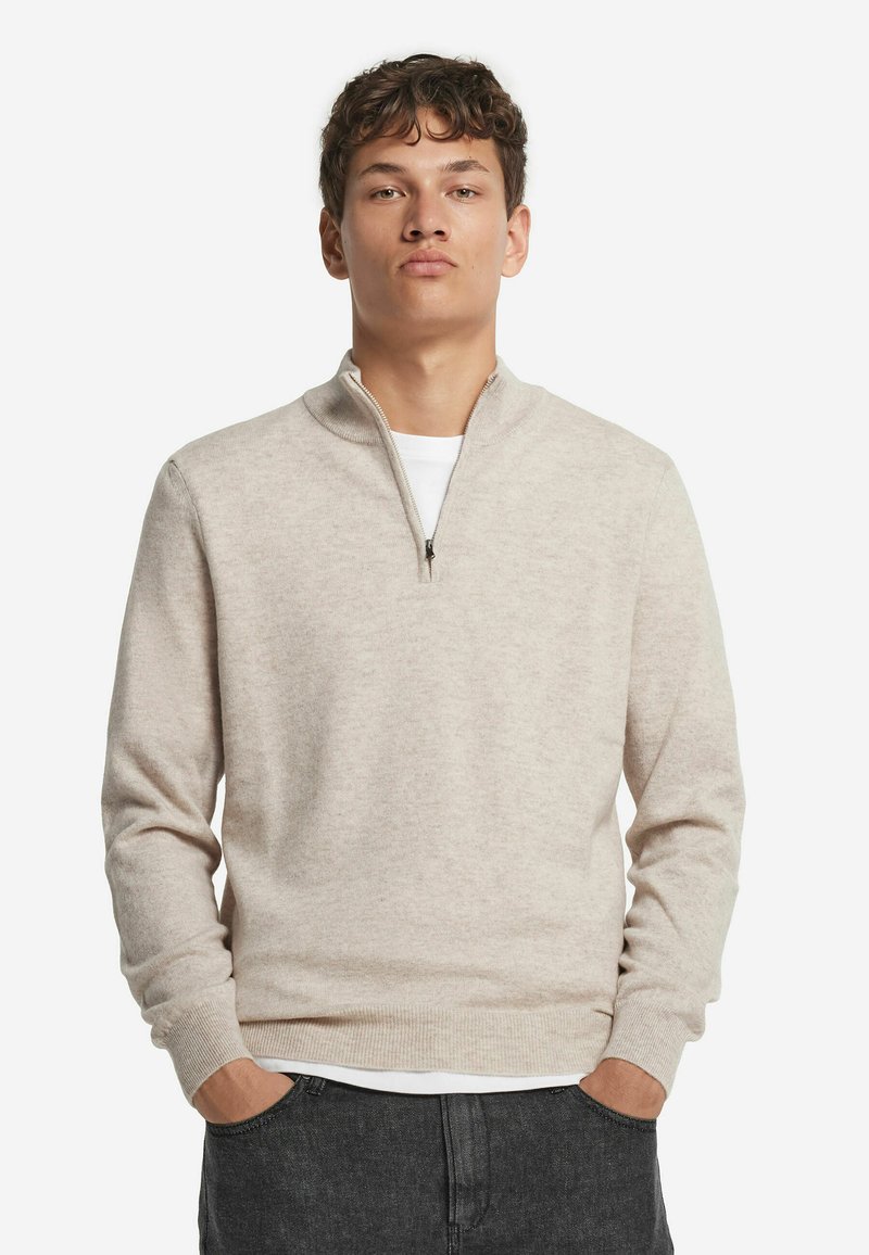 Jasno beżowy sweter z zamkiem przy szyi, wykonany z miękkiego materiału, z długimi rękawami, ściągaczami u rękawów i u dołu, noszony na białej koszuli.