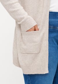 Světlý béžový cardigan s žebrovanými manžetami a přední kapsou. Měkká pletená textura s volným, otevřeným střihem. Nošený přes modré džíny.