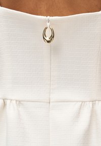 Πίσω όψη λευκού υφασμάτινου ενδύματος με υφή και χρυσό οβάλ φερμουάρ στο κέντρο επάνω.