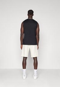 Schwarzes ärmelloses Shirt mit lockerem Schnitt, kombiniert mit hellen Shorts. Weiße Sportsneaker und Socken mit Logo sind getragen.