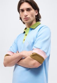 Jeune homme aux cheveux longs jusqu'aux épaules, portant un polo bleu clair avec un col vert et des manches retroussées en rose et marron.