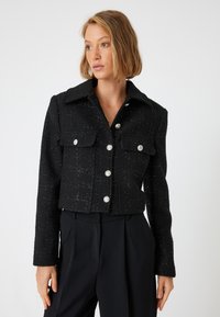 Veste noire coupée en tissu texturé avec un col, deux poches avant et des fermetures à boutons argentés, présentant une coupe ajustée.