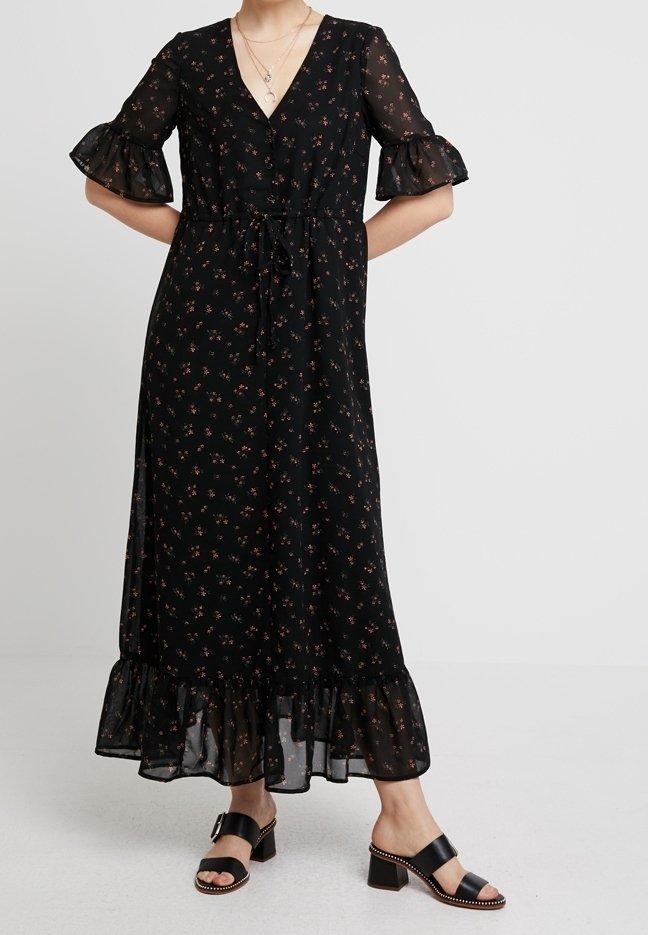 Femme portant une robe midi noire à motifs floraux avec des manches et un ourlet à volants, associée à des sandales à talons carrés noirs avec des bords cloutés.