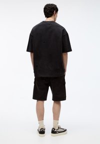 T-shirt noir oversize avec bords effilochés, associée à un short cargo noir. Le modèle porte des chaussettes claires et des baskets noires avec des accents crème.