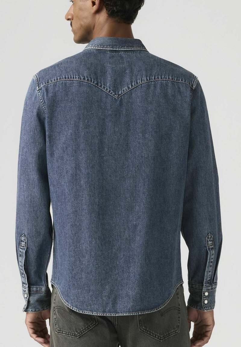 Mann, der ein langärmeliges blaues Jeanshemd und dunkle Hosen trägt, von hinten vor einem schlichten hellen Hintergrund gezeigt.