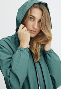 Jachetă verde ușoară cu glugă, manșete elastice și fermoar frontal. Textură netedă și design minimalist.