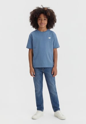 Bambino con capelli ricci in piedi di fronte, indossa una maglietta blu New Balance, jeans blu e sneaker bianche su uno sfondo neutro.