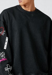 Črna oversized kapuca iz mehkega materiala. Ima rebrast ovratnik ter grafični tekst in dizajne v beli in roza barvi na rokavih.