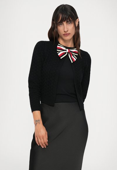 Žena s dlhými tmavými vlasmi má na sebe čierny textúrovaný cardigan, čierny top, čiernu sukňu a veľkú pruhovanú kravatu u goliera.