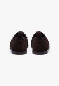 Braune Wildlederschuhe mit gewobener Textur, im Slip-On-Design und einer tiefen schwarzen Gummisohle. Die Rückansicht zeigt die Akzentnähte und Schlaufen.