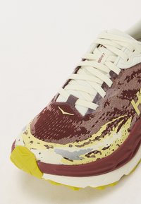 Laufschuh mit einem kastanienbraunen und cremefarbenen Obermaterial, das aus einem strukturierten Stoff besteht, gelben Akzenten und einem markanten Sohlenmuster.