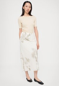 Cremfarbener Kurzarm-Top, kombiniert mit einem weißen Midirock mit floralen Mustern und diagonalen Streifen; Modell trägt schwarze Ballerinas.