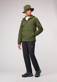 Jachetă verde cu glugă, cu un design cu jumătate de fermoar, pantaloni negri și bocanci de drumeție verzi închise. Dispune de un buzunar frontal și detalii de marcă.