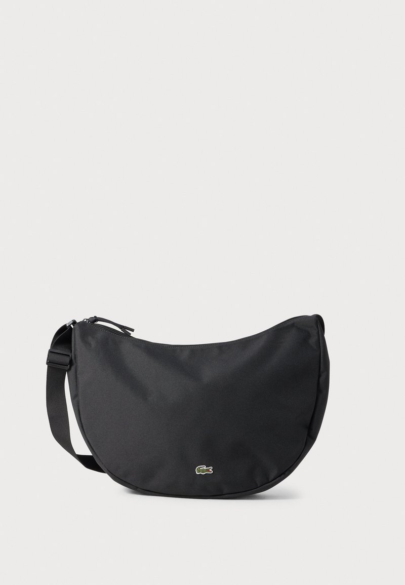 Black fabric crossbody bag with a curved shape, featuring a zip closure and a small green logo on the front. Adjustable shoulder strap.