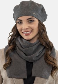 Berretto a maglia grigio con forma rotonda e un piccolo logo metallico, abbinato a una sciarpa grigia coordinata con una morbida texture e un design drappeggiato.