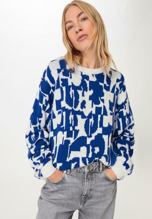 Blauw-wit gebreide trui met een ronde halslijn; heeft losse mouwen en geribde boorden; gecombineerd met grijze jeans.