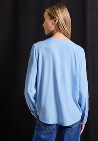 Blouse bleu clair en tissu lisse, avec des manches longues, une coupe décontractée et un ourlet arrondi, associée à un jean bleu.