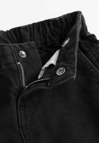 Zbliżenie na czarne spodnie z elastycznym paskiem, częściowo rozpięty zamek błyskawiczny i metalowe napy w okolicy pasa.