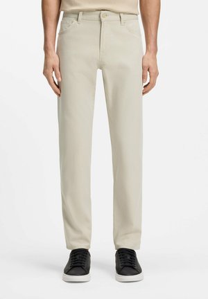 Vista frontale di una persona che indossa pantaloni beige a gamba dritta e sneakers nere con suola bianca su uno sfondo semplice.