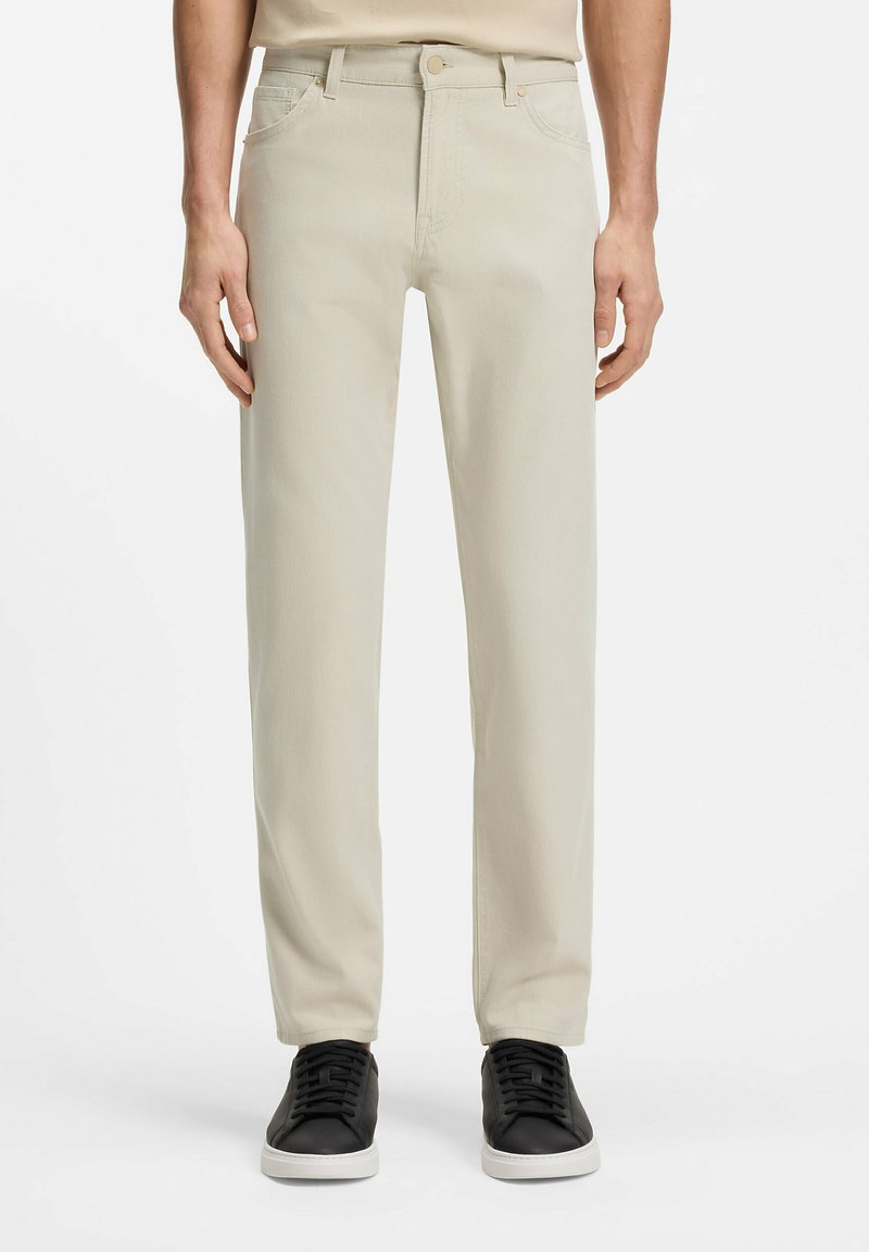 Vue de face d'une personne portant un pantalon beige à jambes droites et des baskets noires à semelles blanches sur un fond uni.