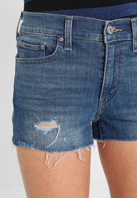 Shorts de mezclilla azul con un diseño desgastado, bordes deshilachados y dos bolsillos frontales. La tela es suave con un acabado deslavado.
