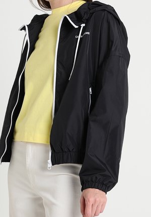 Person, die eine schwarze Kapuzenjacke mit Reißverschluss über einem gelben Shirt und weißen Hosen trägt, vor einem schlichten Hintergrund steht.