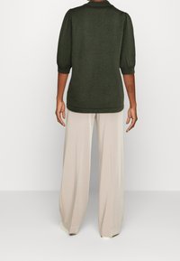 Top en maille côtelée vert foncé avec des manches bouffantes trois quarts, porté avec un pantalon large beige clair. Tissu lisse et coupe décontractée.