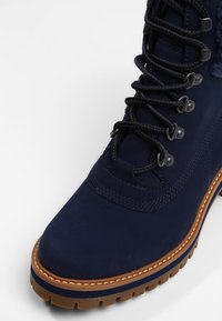 Botte en daim bleu marine avec semelle épaisse en caoutchouc, dotée d'œillets en métal, de lacets contrastants et de coutures texturées le long de la tige et de la semelle.