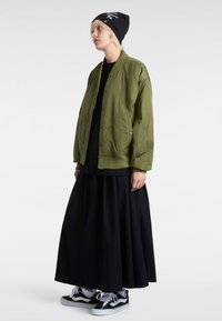 Olive green quilted bomber jacket, black maxi skirt, and black beanie. White and black sneakers with contrasting stripes. Minimalistic design.