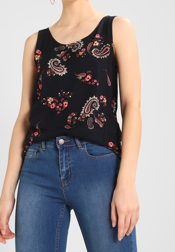 Femme portant un haut noir sans manches avec des motifs floraux cachemire rouges et beiges, accompagné d'un jean bleu taille haute.